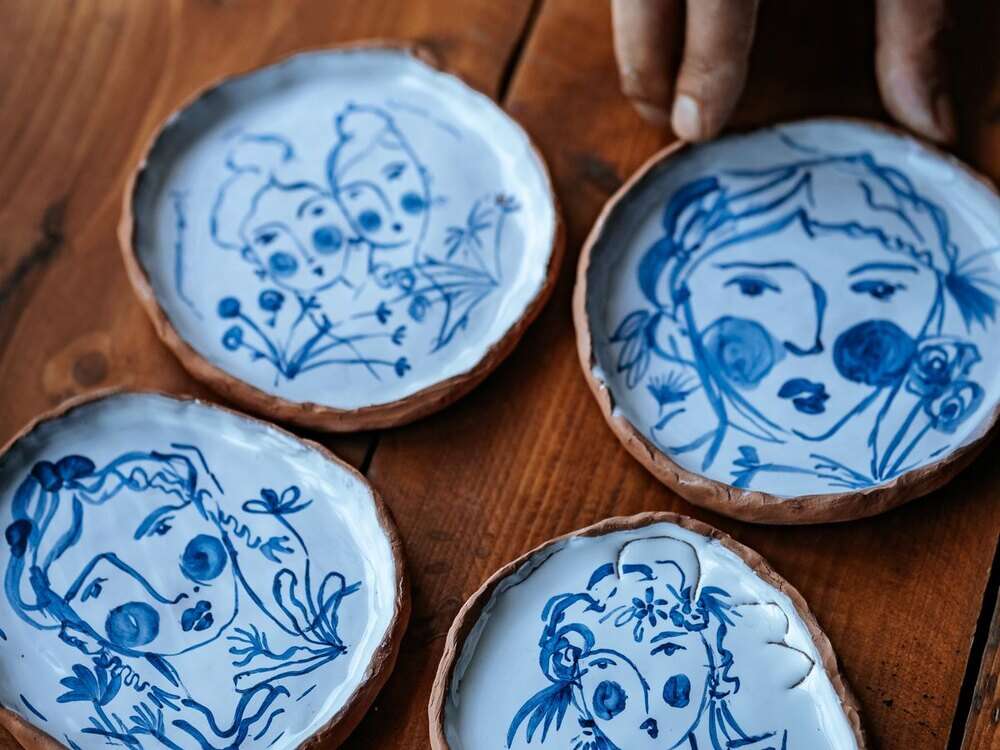 Pratos pequenos de cerâmica pintados à mão com desenhos em azul, sobre mesa de madeira, com a mão do artista ao lado